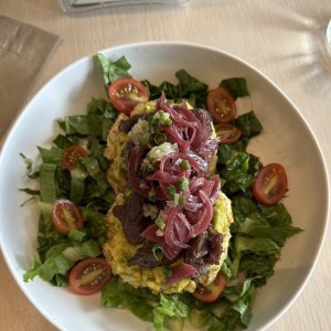 Dos patacones asados con aceite de oliva, cubiertos de guacamole, entraña, chimichurri y cebolla encurtida, sobre ensalada de lechuga y tomate con aderezo de limón